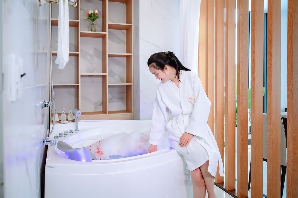 A woman in a white robe in a modern bathroom with a marble vanity, a shower, and a wooden shelf with flowers.