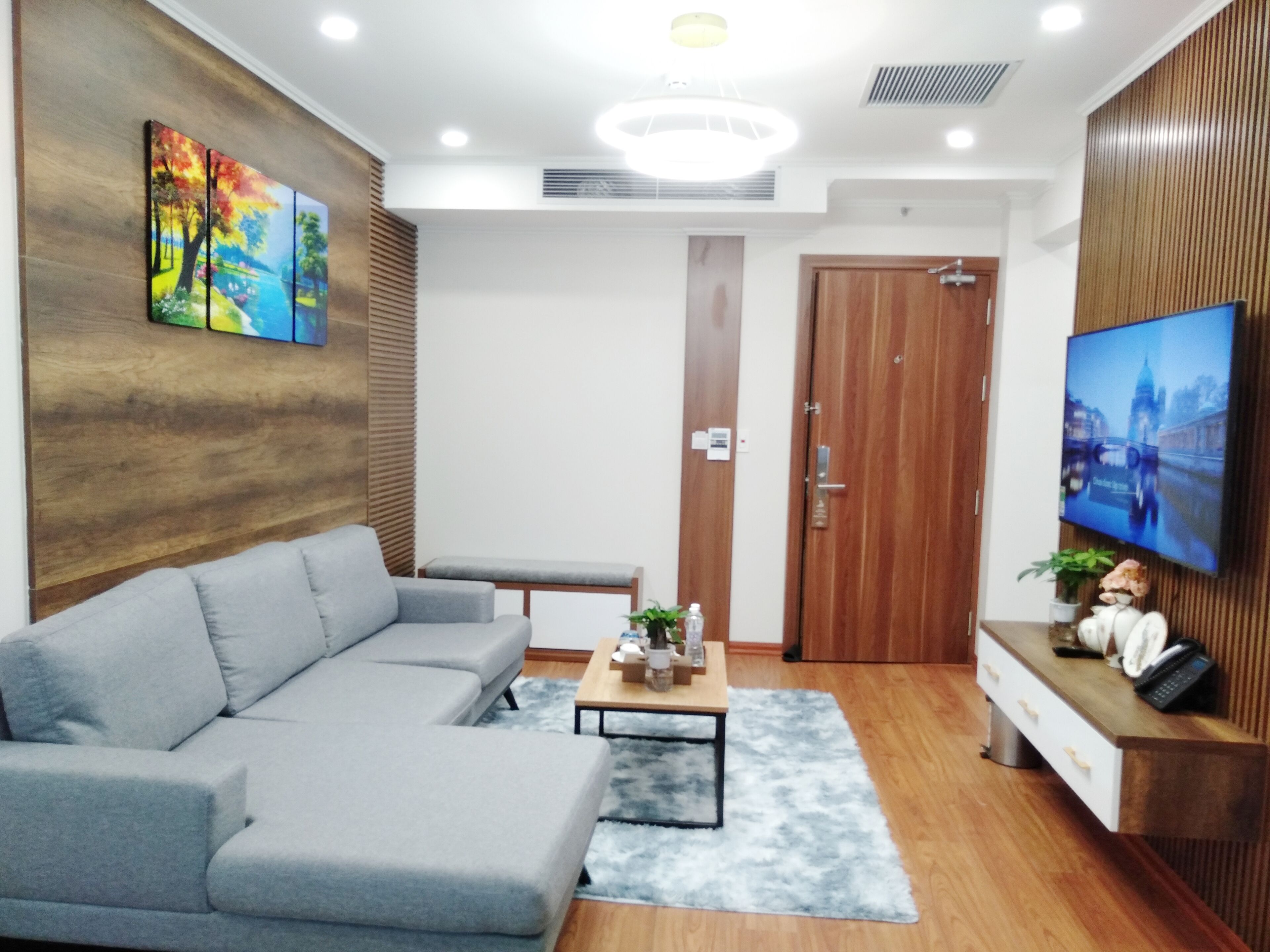 A modern living room with a grey sectional sofa, a wooden accent wall, a flat-screen TV, and a wooden reception desk.