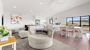 Un salon moderne avec un canapé rond de couleur grise, un coin repas avec des chaises blanches et une cuisine en arrière-plan.