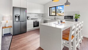 Une cuisine moderne avec des armoires blanches, des appareils en acier inoxydable, un îlot central doté de sièges et une vue sur une maison par une fenêtre.