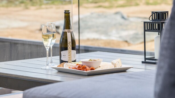 Une table avec une bouteille de vin Brut Handon, deux verres de vin blanc, un bol de nourriture et une assiette avec du fromage et du pain.