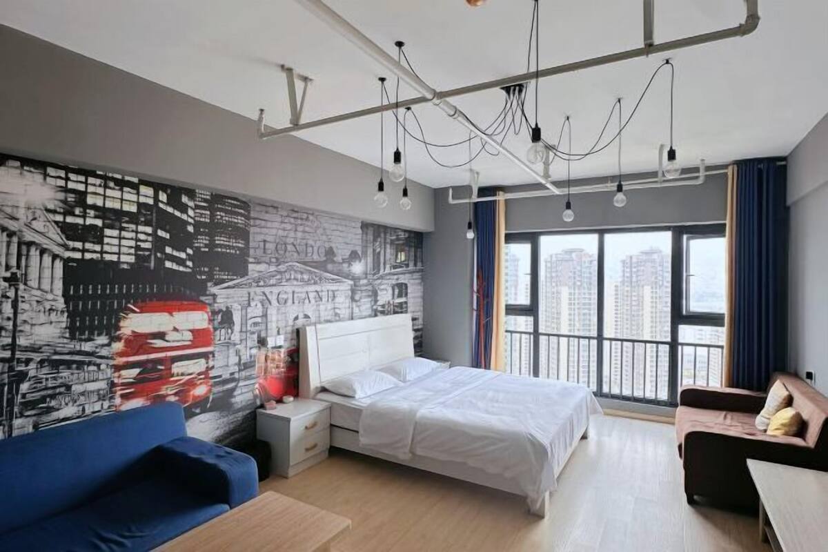 A modern hotel room with a large bed, a blue sofa, a small table, and a cityscape mural on the wall.