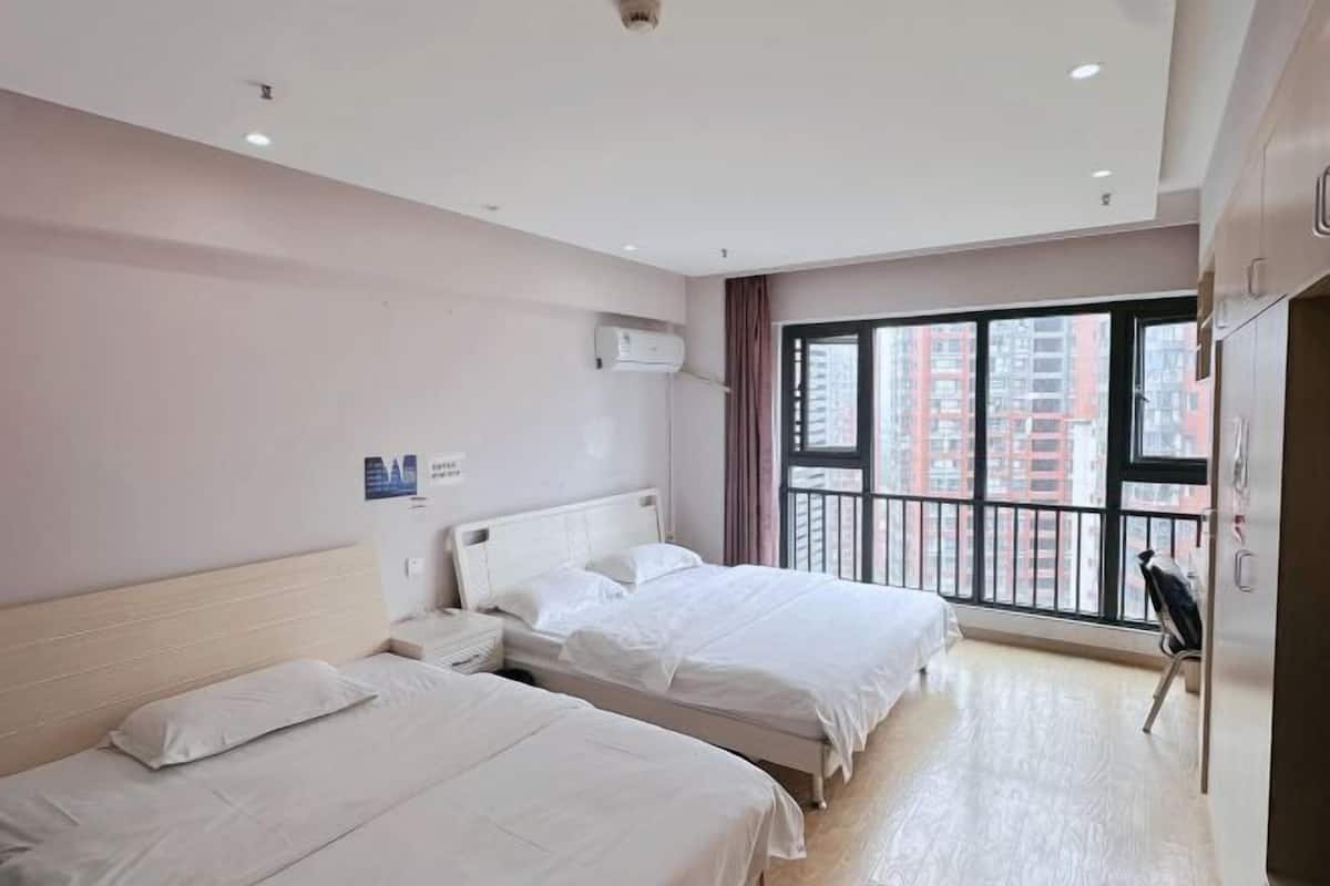 A hotel room with two beds, a window with a view of buildings, a ceiling fan, and a wall-mounted air conditioner.