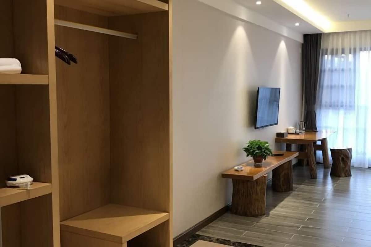 A modern hotel room with a wooden wardrobe, a small table with a plant, a TV mounted on the wall, and a dining area with a wooden table and chairs.