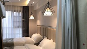 Ein Hotelzimmer mit einem Bett, zwei Pendelleuchten, einem Fenster mit Vorhängen und einem an der Wand montierten Kopfteil.