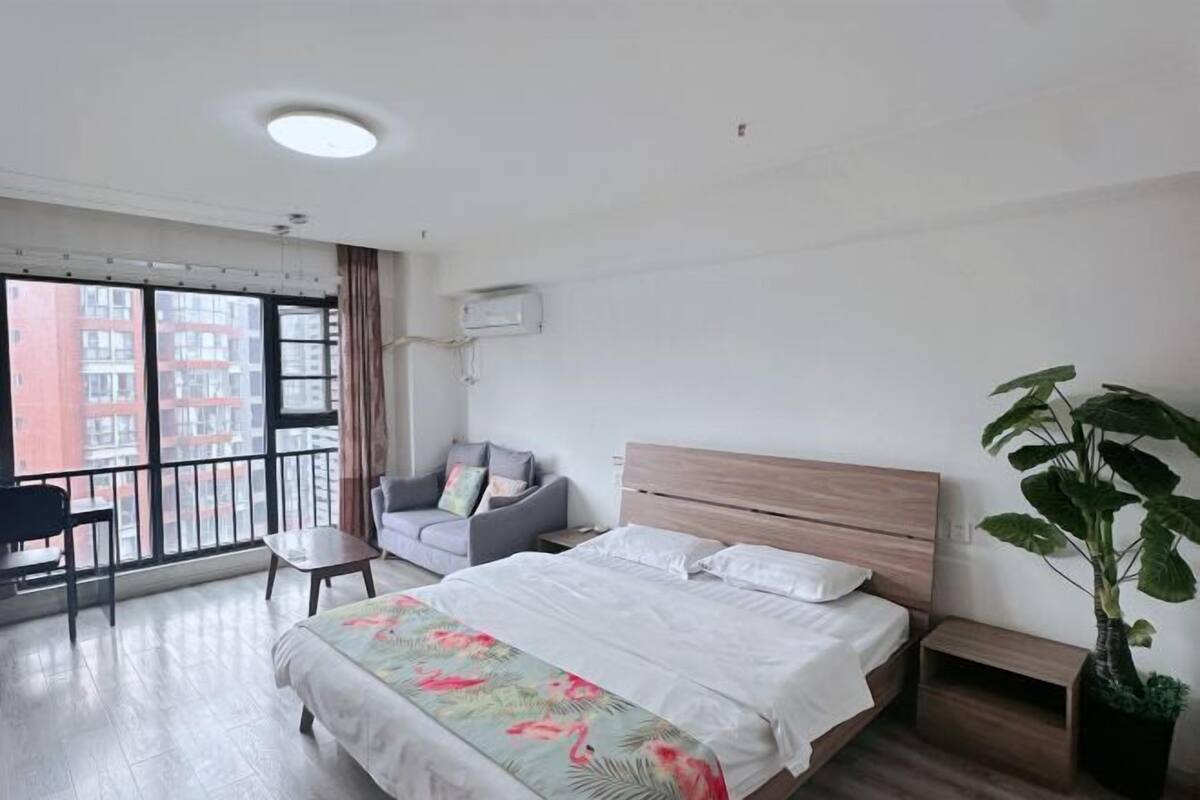 A hotel room with a bed, a chair, a small table, a potted plant, and a balcony with a view of buildings.