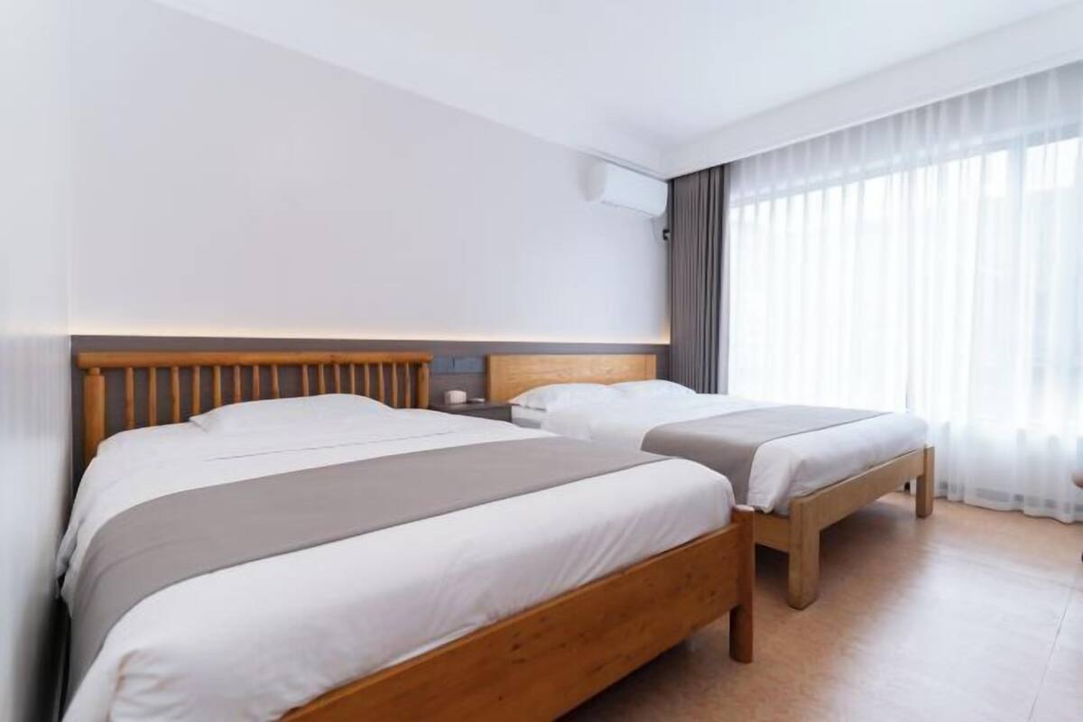 A hotel room with two beds, a wooden headboard, a bedside table, and a large window with curtains.