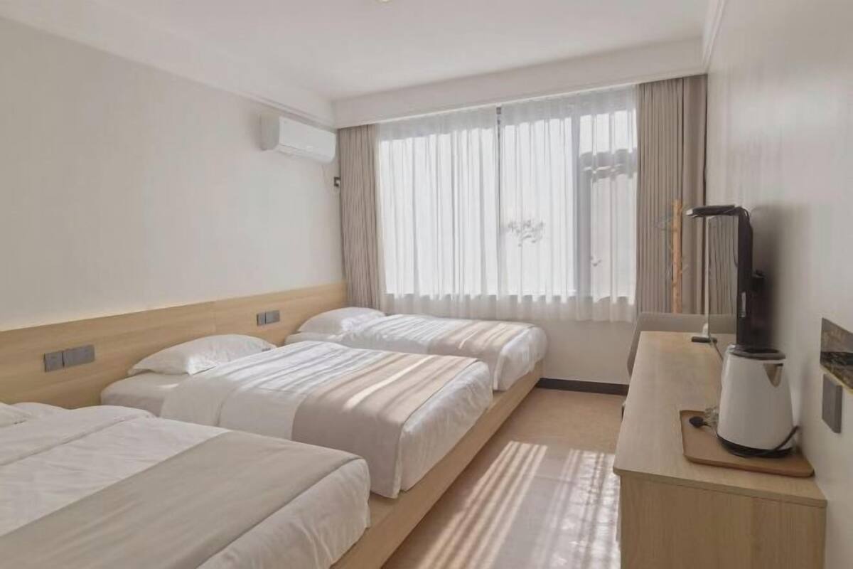 A hotel room with two beds, a desk with a coffee maker, a large window with curtains, and an air conditioning unit.