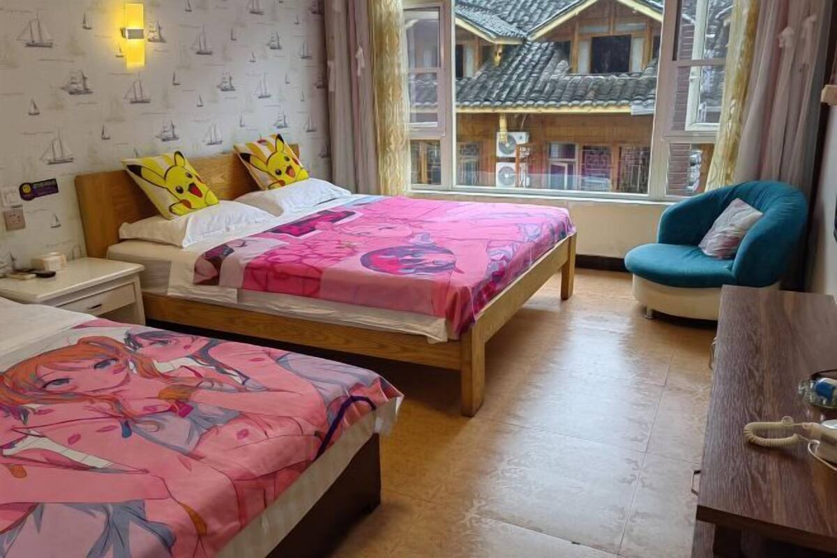A hotel room with two beds, a chair, a wooden desk, and a window with a view of a traditional building.