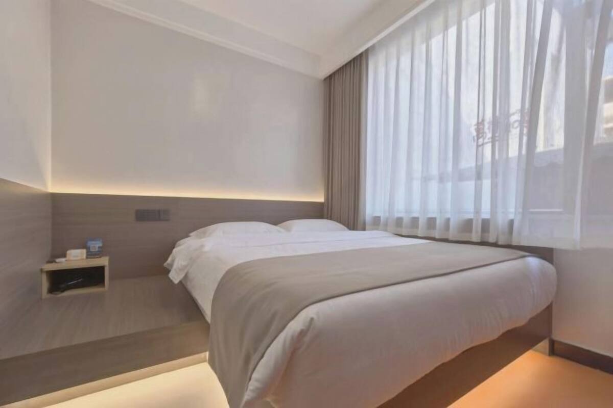 A modern hotel room with a bed, bedside table, and a window with sheer curtains.