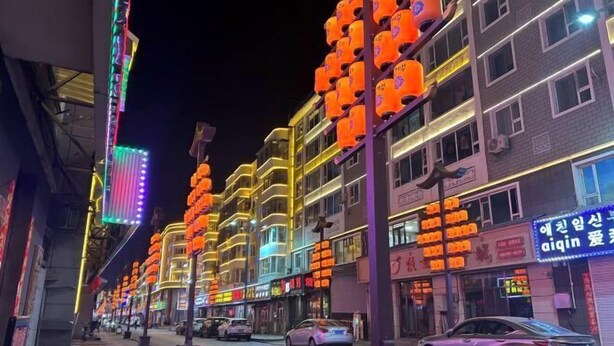 A brightly lit street with multi-story buildings, illuminated signs, and parked cars. - Jiang Zhi Yuan Inn (Changbai County)