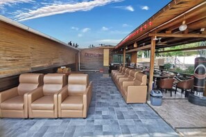 A covered outdoor seating area with leather chairs and tables, a wooden wall, and a clear sky.