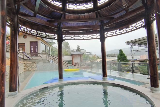 A hot spring area with a wooden gazebo structure, a pool, and a view of a building and landscape.