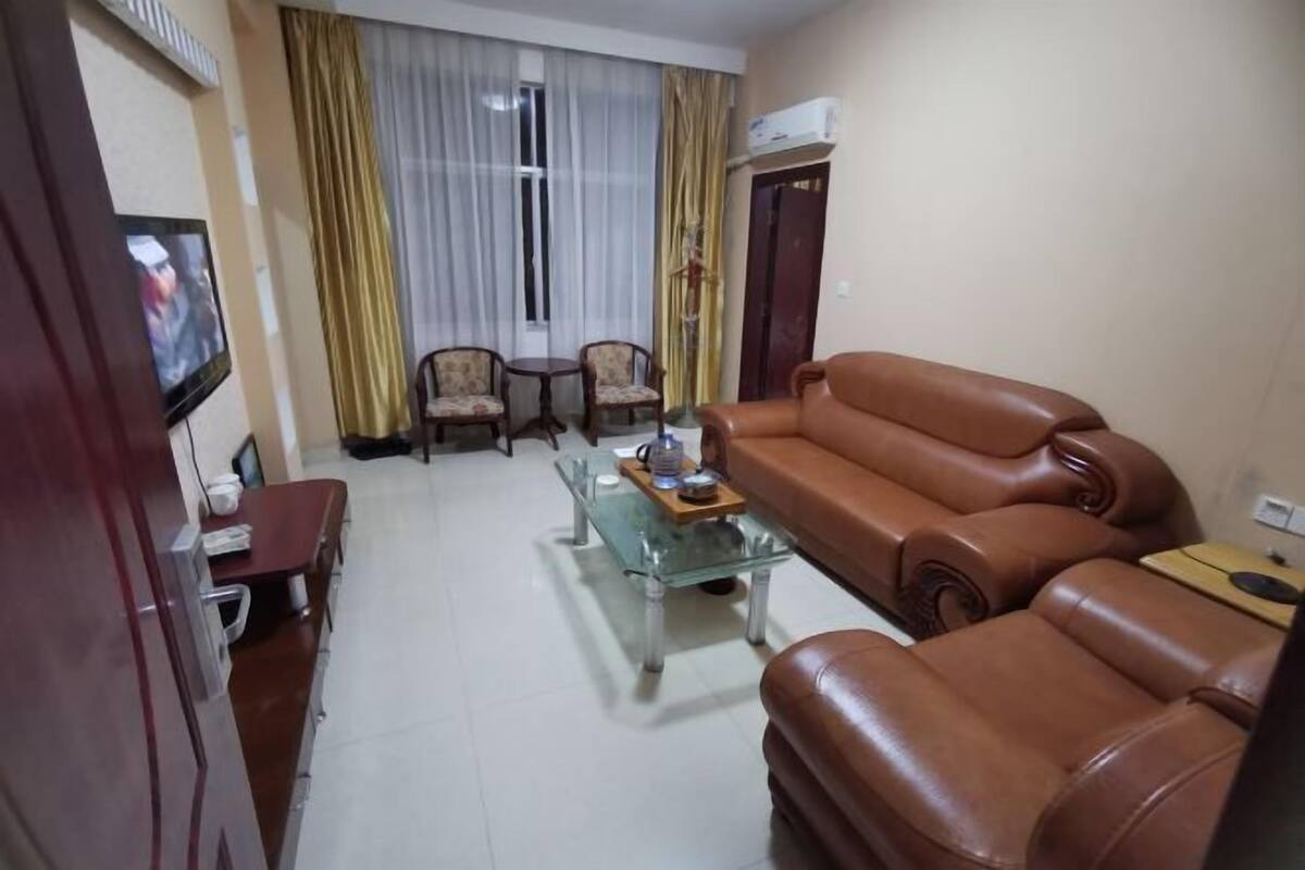 A living room with a brown leather sofa, a glass coffee table, two armchairs, a TV mounted on the wall, and a door with an air conditioning unit.
