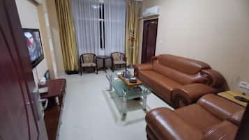Una sala de estar con un sofá de cuero marrón, una mesa de centro de vidrio, dos sillones, un televisor montado en la pared y una puerta con un equipo de aire acondicionado.