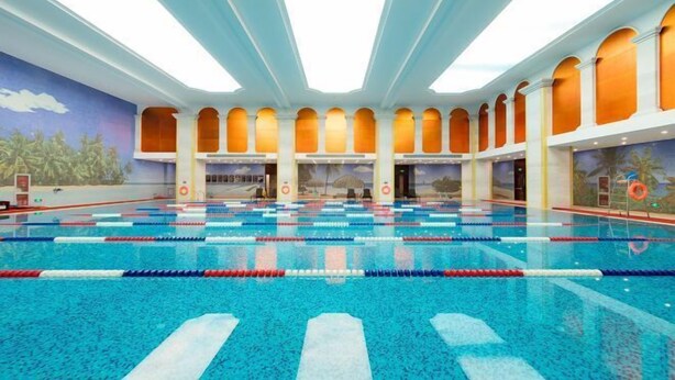 Una piscina interior con varias pistas, un techo alto con tragaluces y un mural de paisaje tropical en las paredes.
