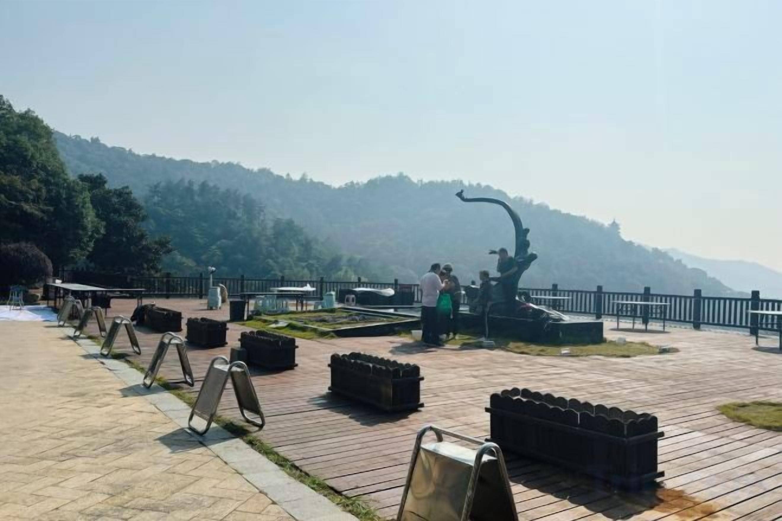 A paved walkway with benches and a sculpture, surrounded by a fence and trees, leading to a misty hill.