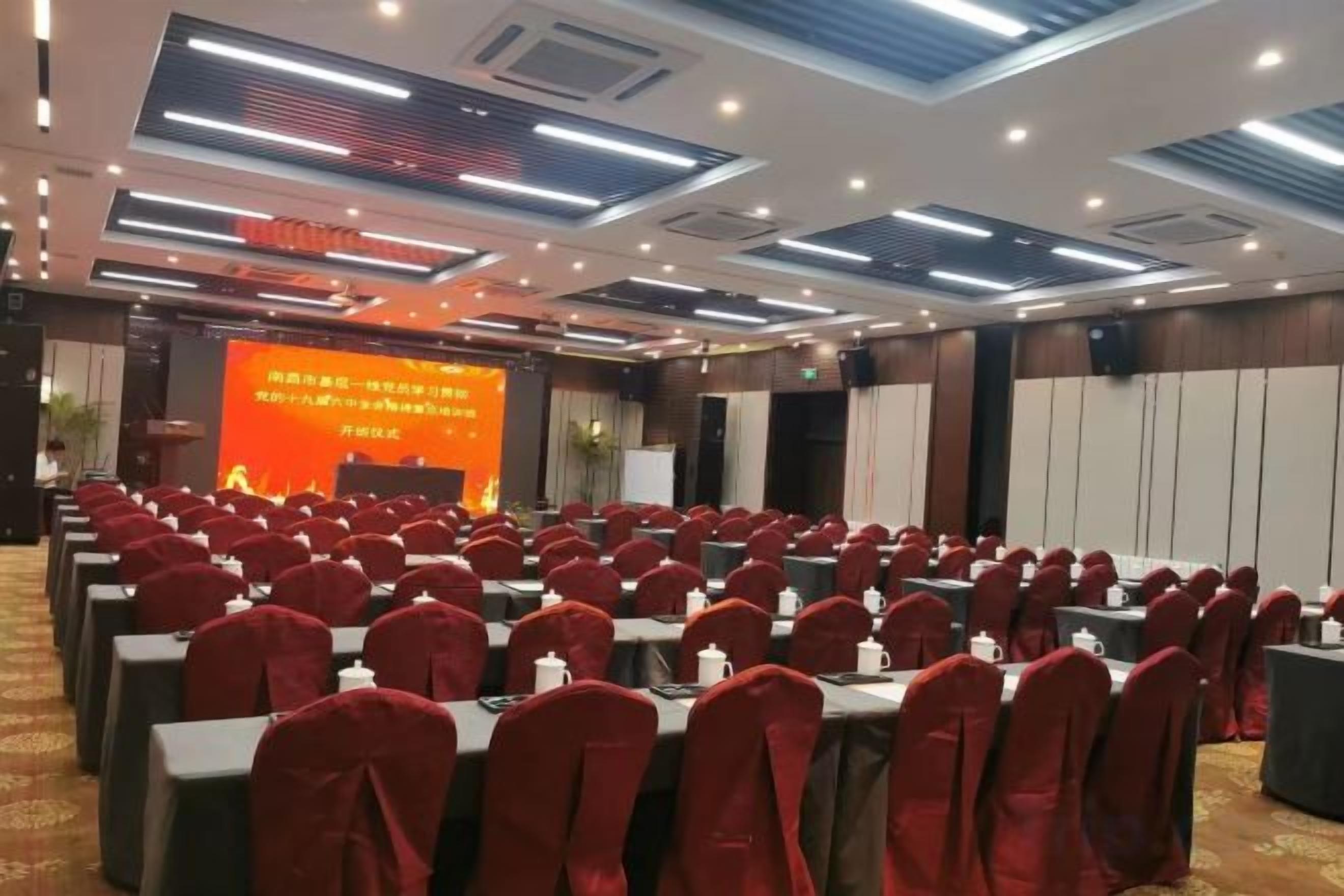 Une salle de conférence avec des rangées de tables et de chaises, une scène dotée d’un écran de présentation et un plafond équipé d’un éclairage encastré.