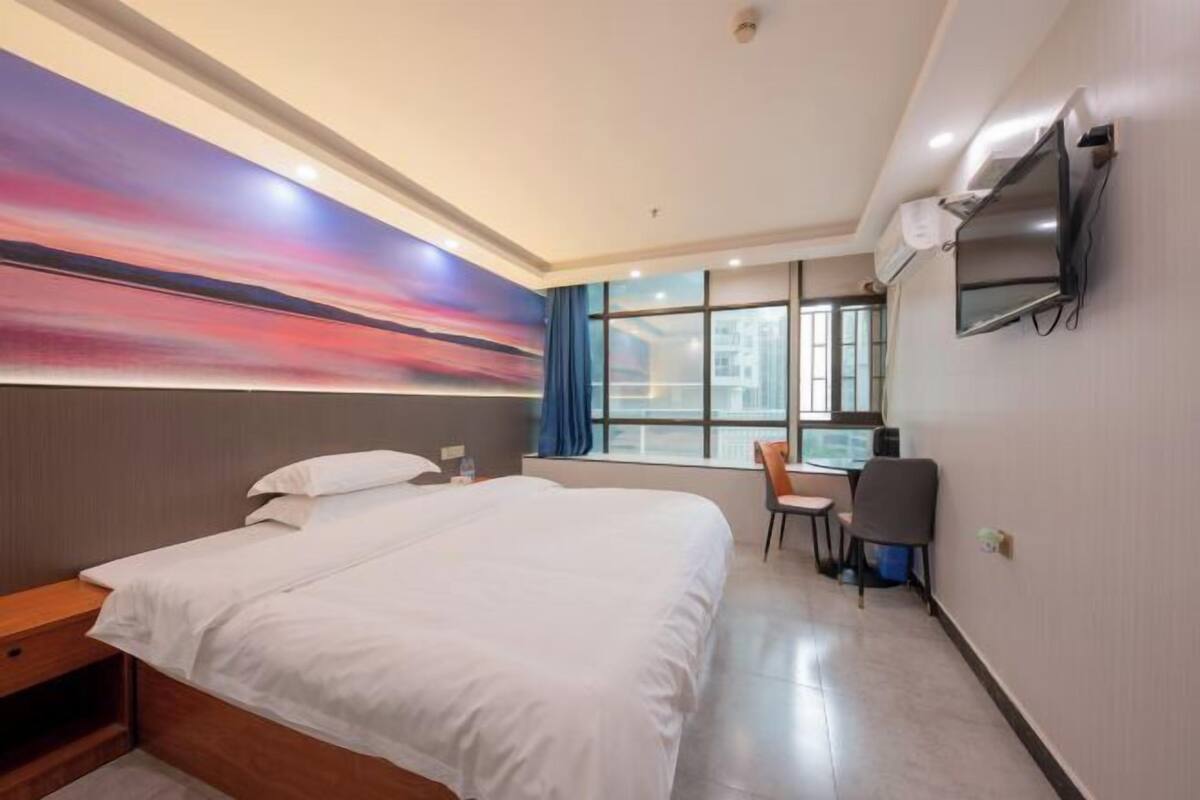A hotel room with a large bed, a desk with a chair, a TV mounted on the wall, and a window with curtains.