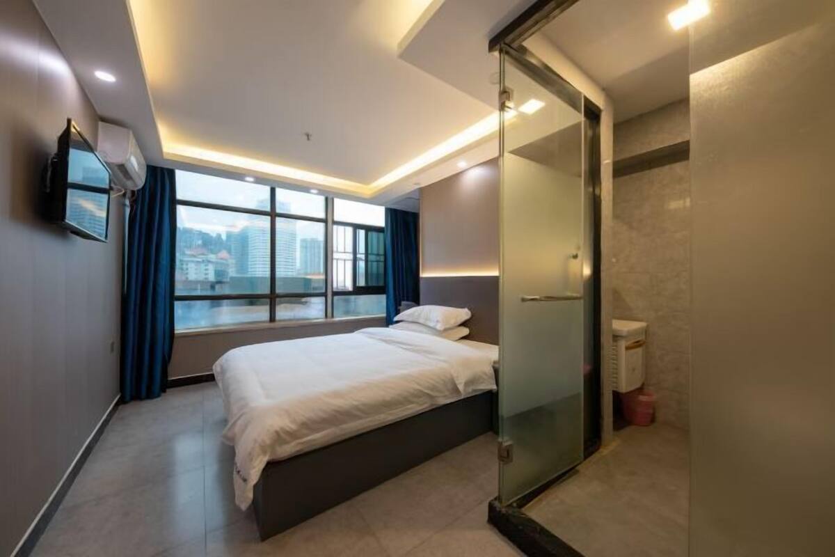 A modern hotel room with a large bed, a glass shower enclosure, a television mounted on the wall, and a view of the cityscape through the window.