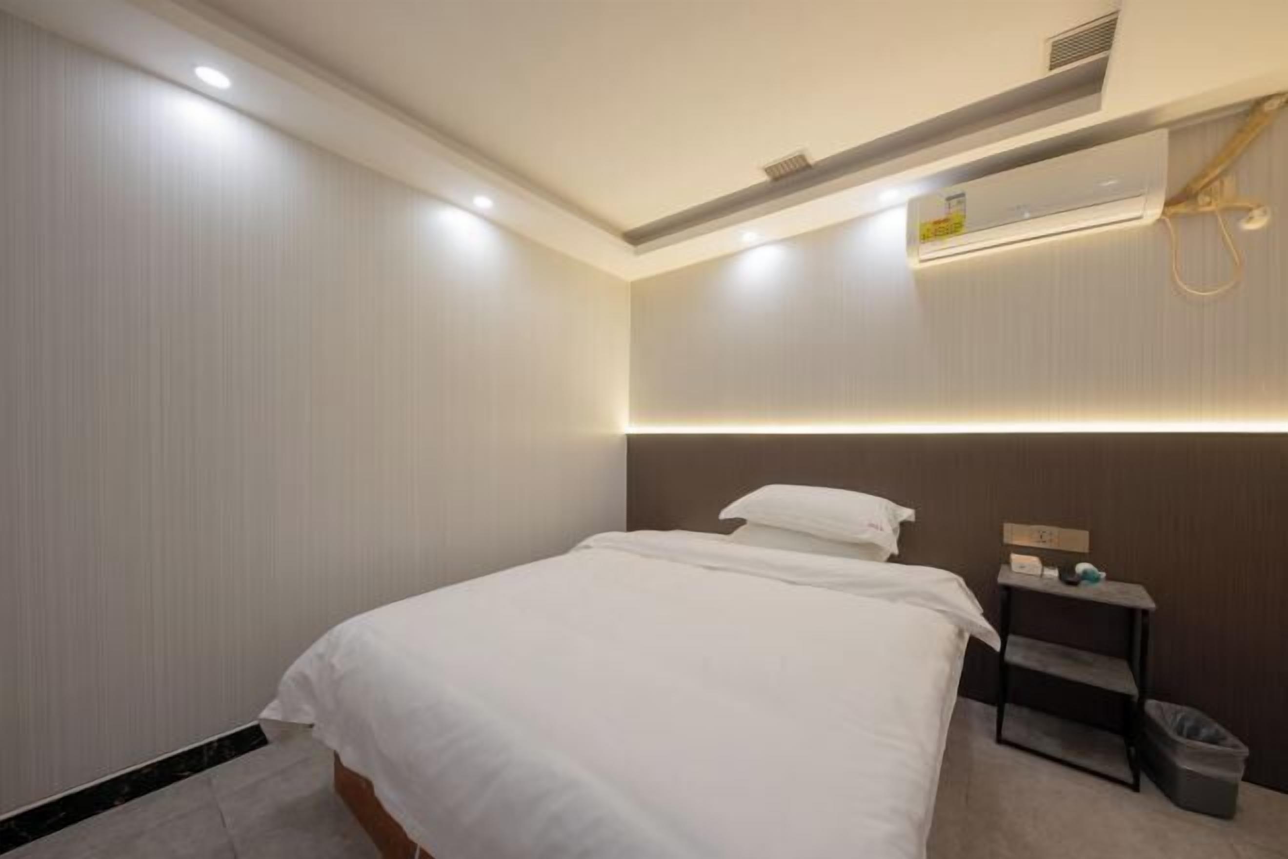 Een hotelkamer met een bed, nachtkastje, prullenbak en een wandairconditioner.