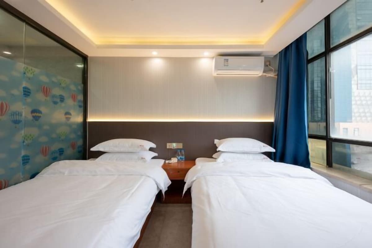 A hotel room with two beds, a nightstand, an air conditioning unit, and a window with curtains.