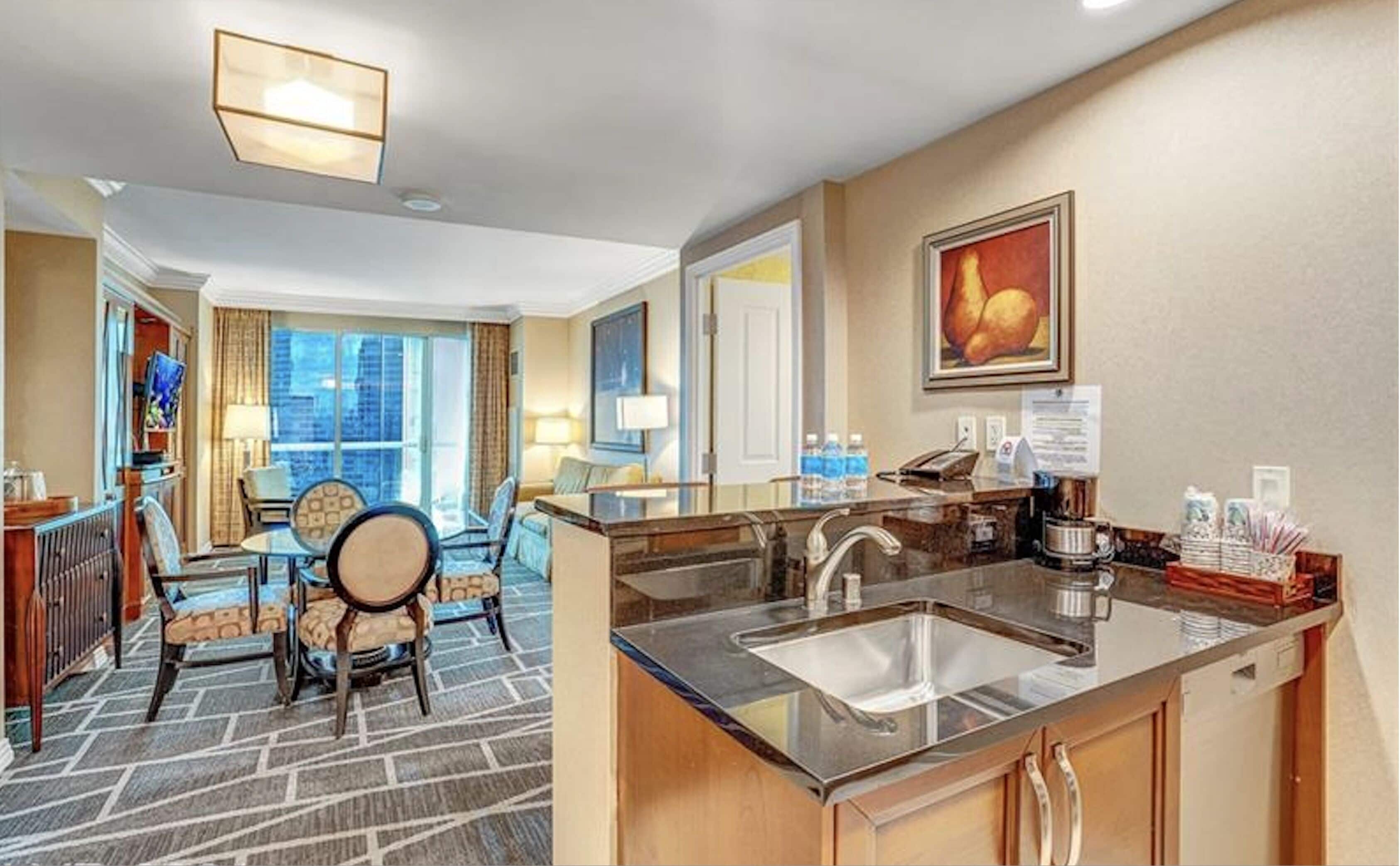 A hotel room with a kitchenette, a dining area with a table and chairs, a large window, and a painting on the wall.
