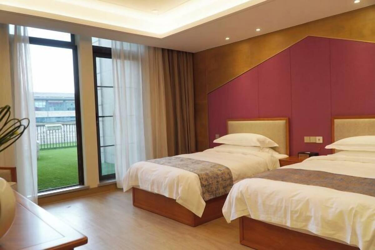 A hotel room with two beds, a large window with curtains, a wooden floor, and a view of a green area outside.