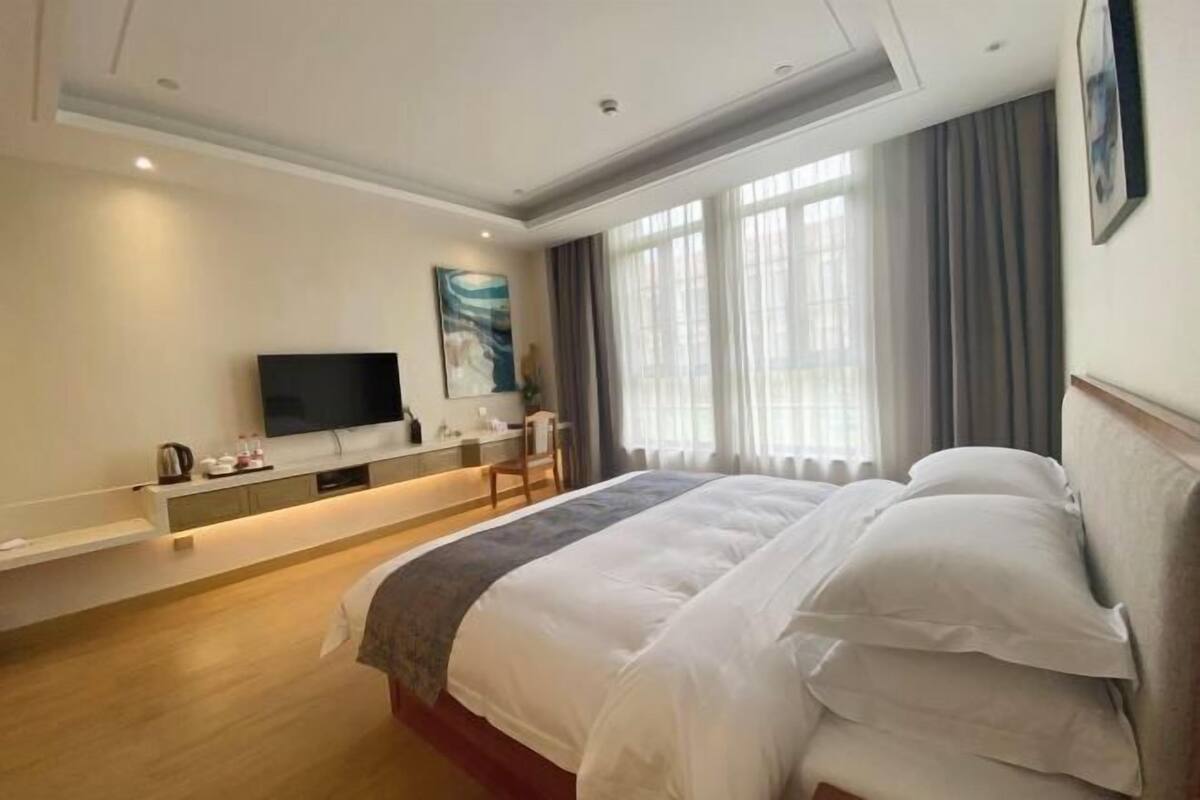 A hotel room with a large bed, a flat-screen TV mounted on the wall, a bedside table with a lamp, and a window with curtains.