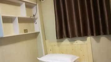 A hotel room with a wooden headboard, a bed with white bedding, a shelf with a few items, and a brown curtain.
