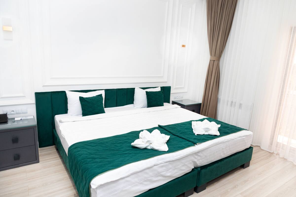 A neatly made bed with green bedding and white pillows, a bedside table with a lamp, and a window with curtains.