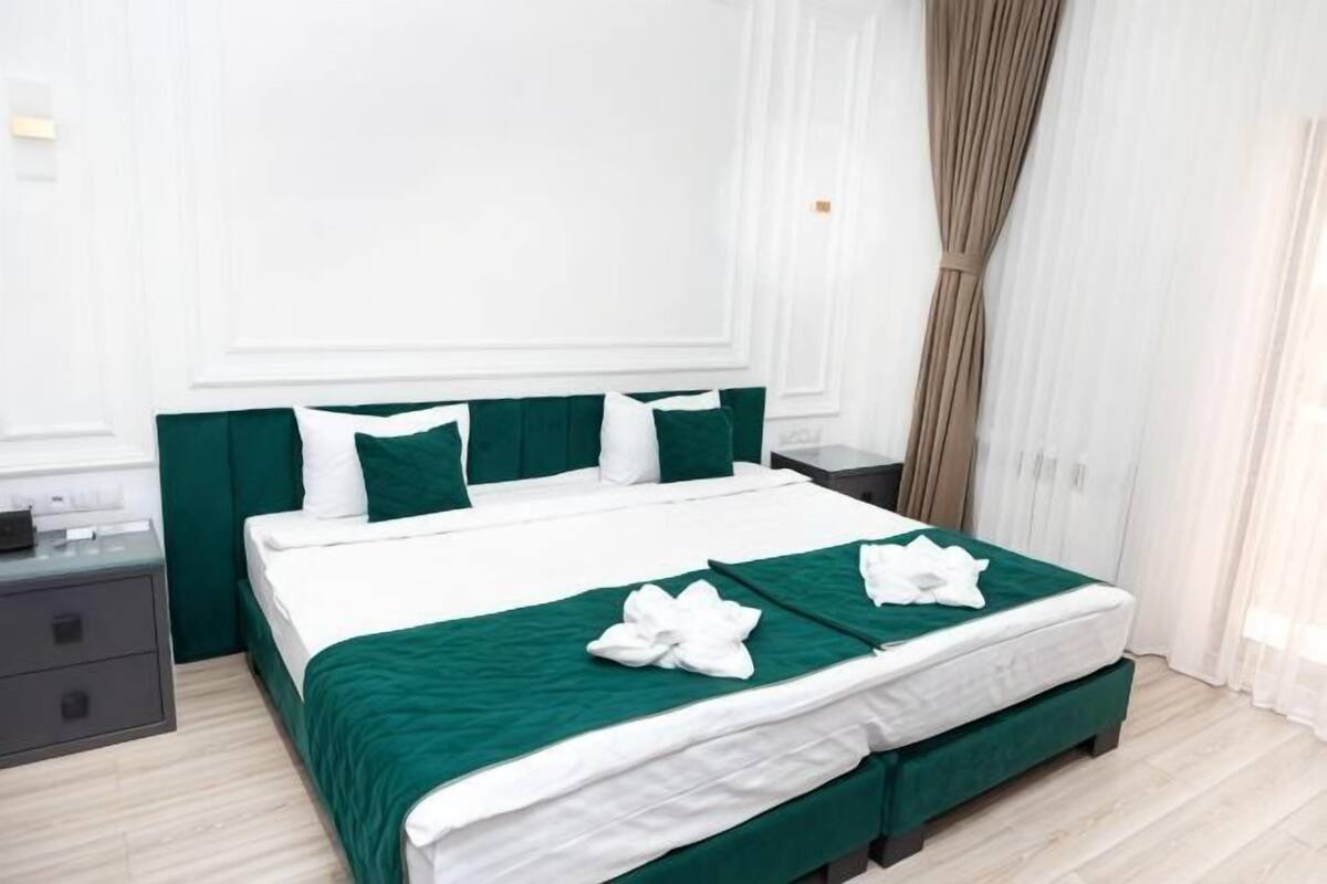 A neatly made bed with green bedding, a bedside table, and a window with curtains.