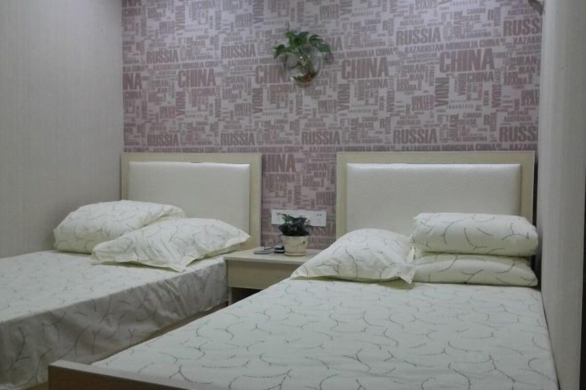 A hotel room with two single beds, a small bedside table with a plant, and a wall with a word cloud design.