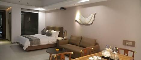 A modern hotel room with a bed, a sofa, a dining table with chairs, and a wall-mounted light fixture.