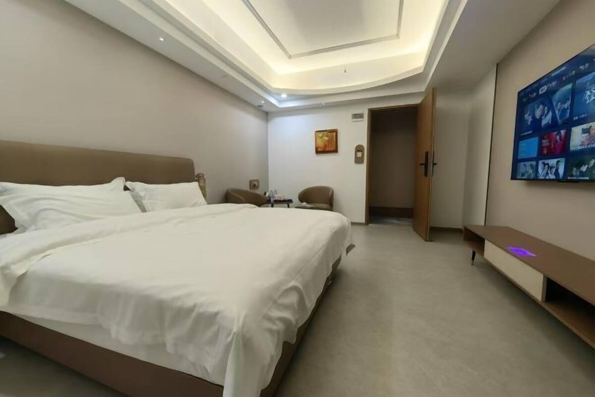 A hotel room with a large bed, a desk, a chair, a TV mounted on the wall, and a door leading to another room.
