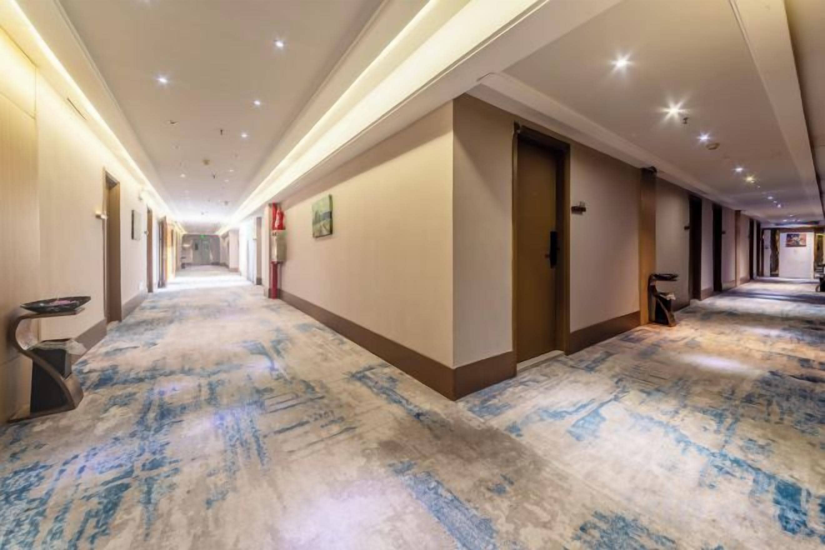 Un couloir d’hôtel avec un sol recouvert de moquette, des portes de chaque côté et un éclairage au plafond.