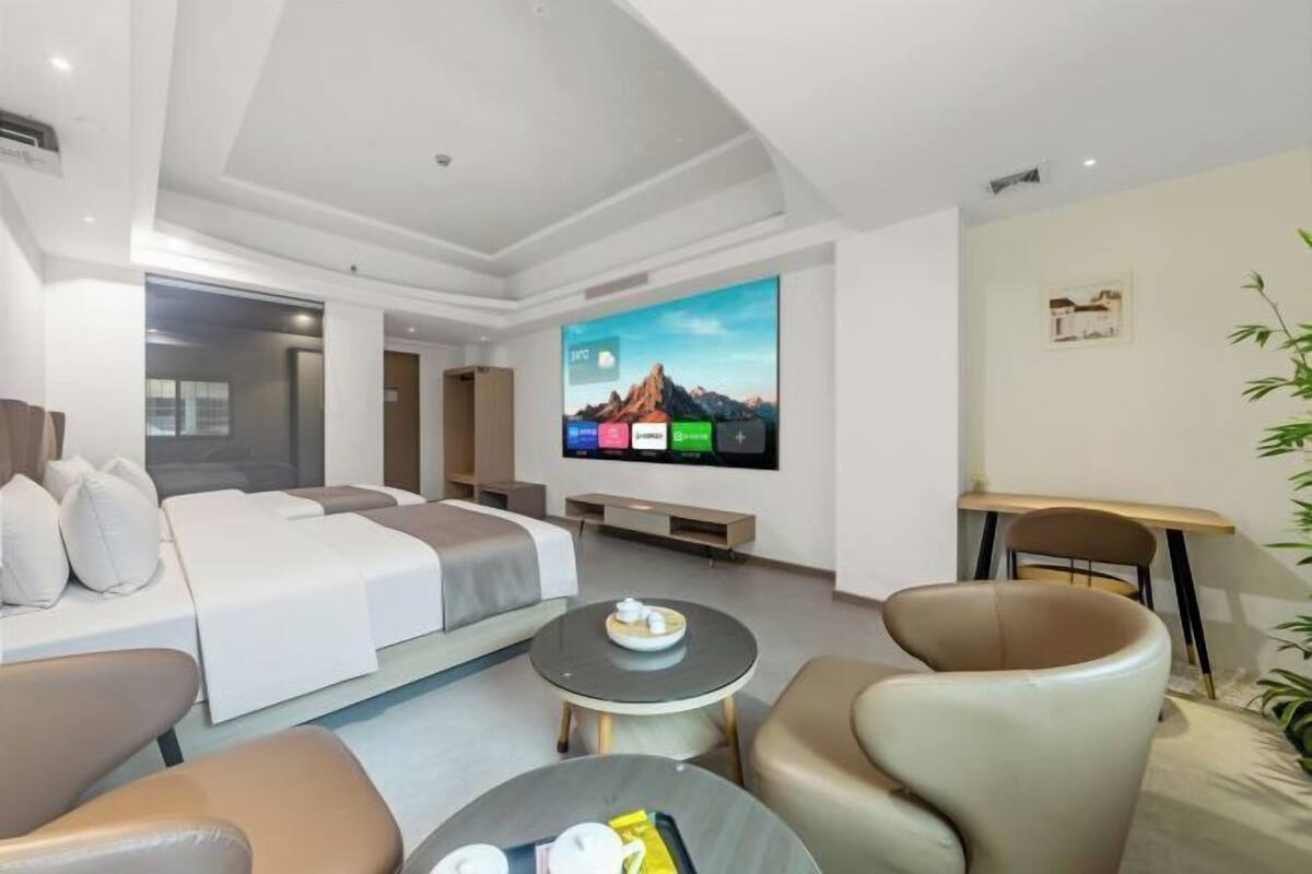 A modern hotel room with a bed, two armchairs, a round coffee table, a TV, and a small table with a plant.