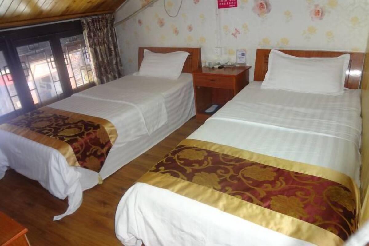 A room with two beds, each with a wooden headboard and white bedding. There is a wooden nightstand between the beds, and a small table with a lamp. The room has a wooden floor and a window with curtains.
