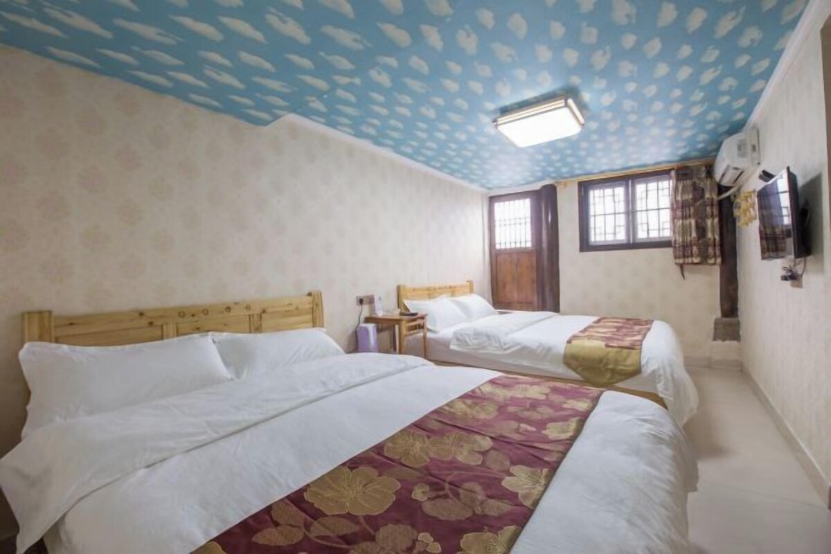 A room with two beds, a wooden headboard, a window with curtains, and a wall-mounted TV.