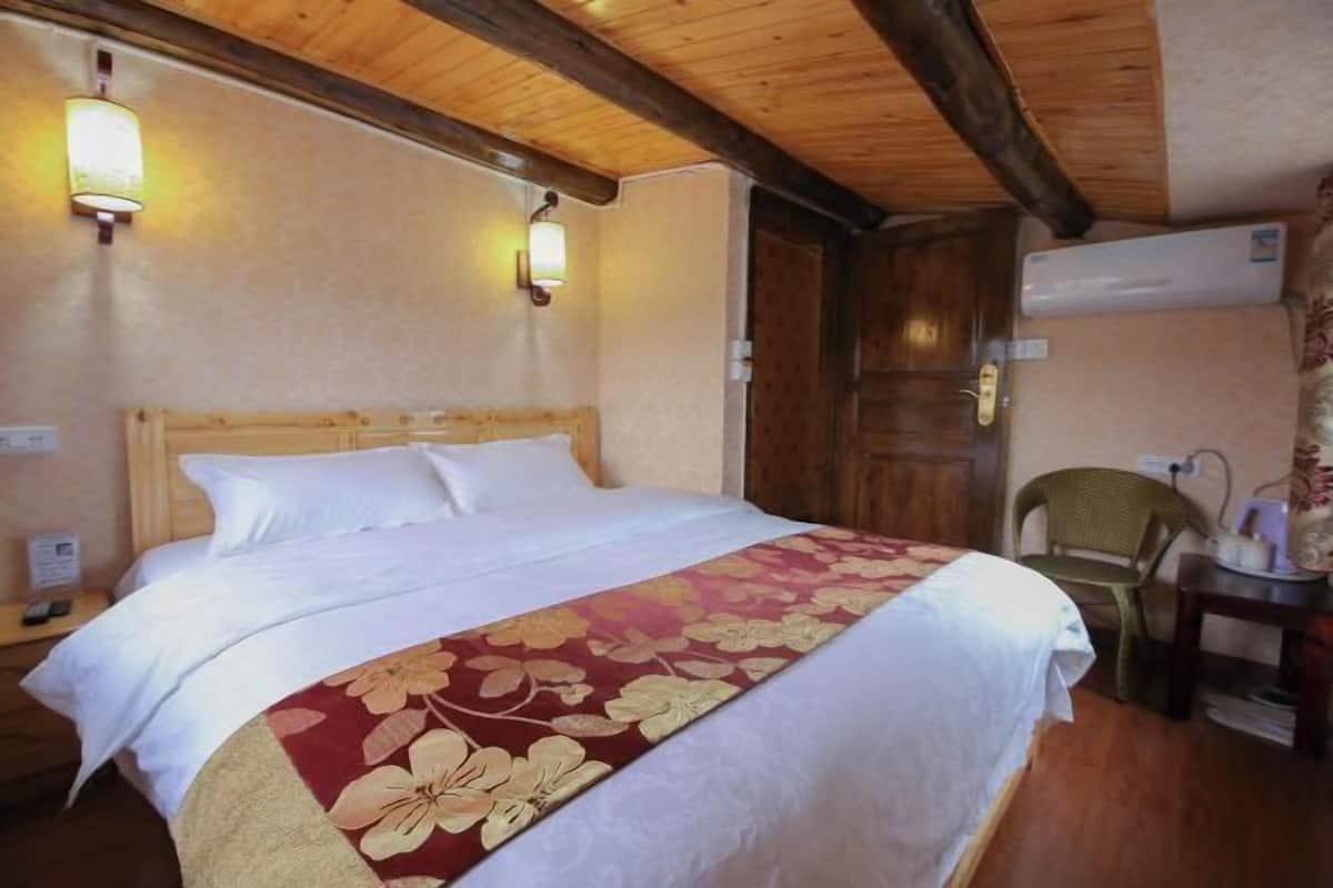 A hotel room with a wooden ceiling, a bed with a floral bedspread, two wall-mounted lights, a wooden door, and a chair.