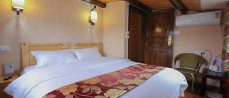 A hotel room with a wooden ceiling, a bed with a floral bedspread, two wall-mounted lights, a wooden door, and a chair.