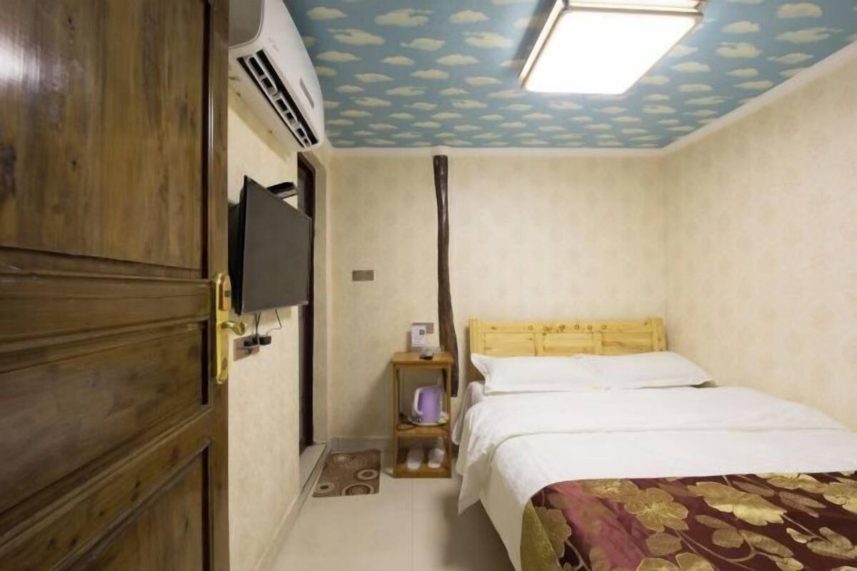 A room with a wooden door, a bed with a floral bedspread, a nightstand with a lamp, and a ceiling with a cloud pattern.