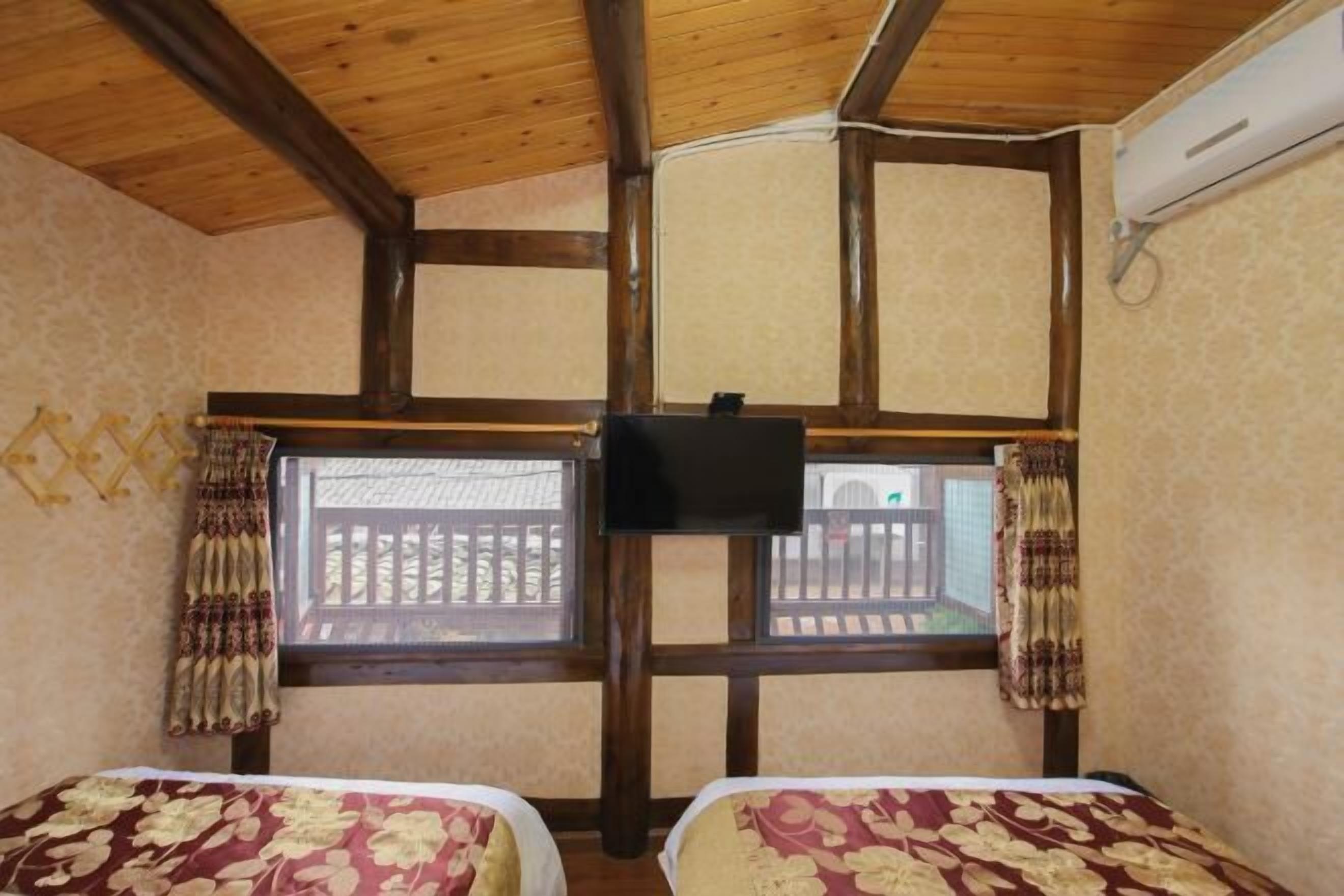A room with two beds, wooden ceiling beams, a television mounted on the wall, and a window with a view of a balcony.