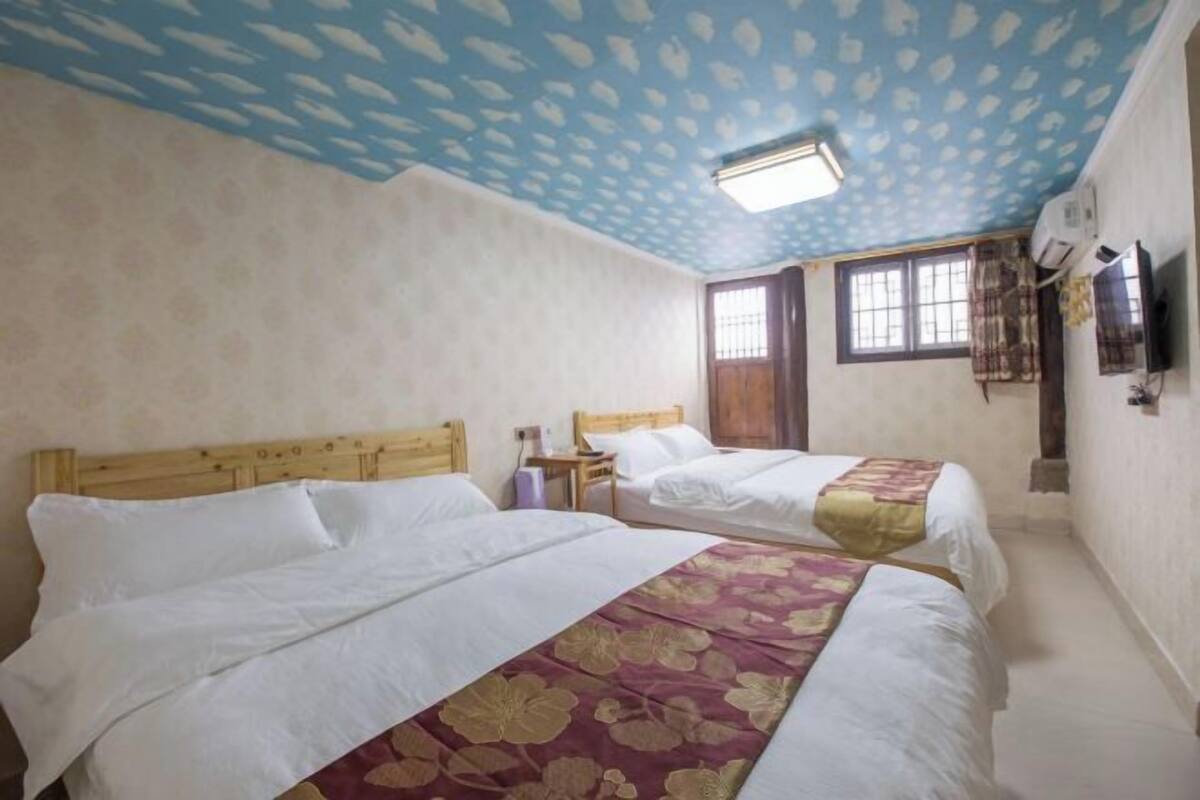 A hotel room with two beds, a wooden headboard, a window with curtains, a wall-mounted TV, and an air conditioning unit.