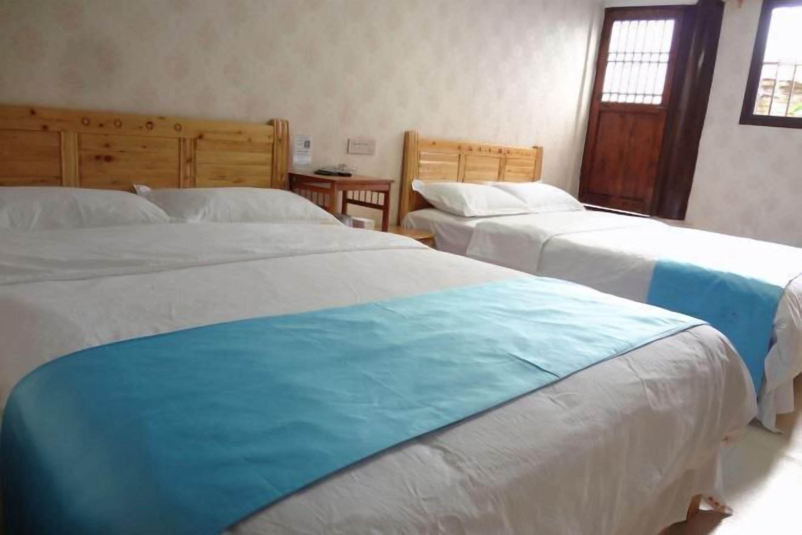 A room with two beds, each with wooden headboards and white bedding. There is a small wooden table and a window with a wooden door.