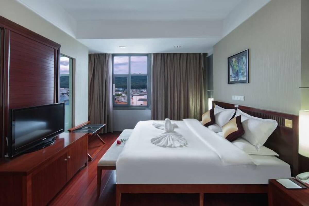 A hotel room with a large bed, a TV on a wooden cabinet, a window with curtains, and a painting on the wall.