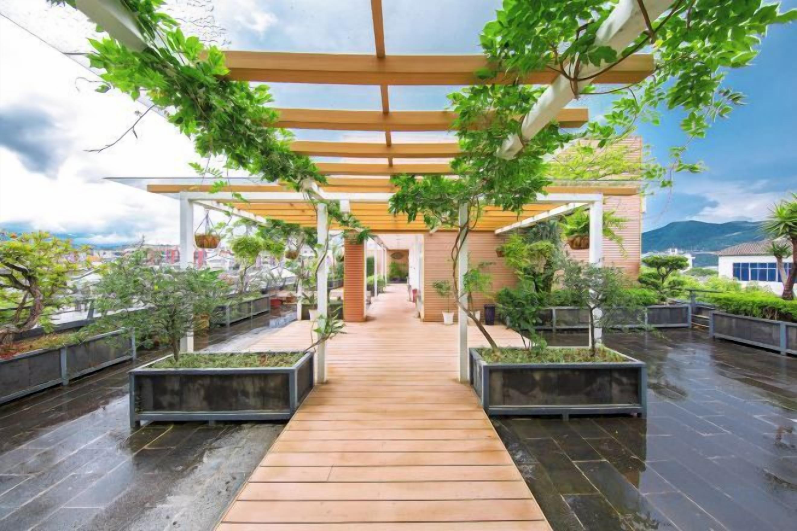 Une allée couverte, dotée d’un plancher en bois, mène à un bâtiment, lequel est entouré de jardinières et d’arbres.