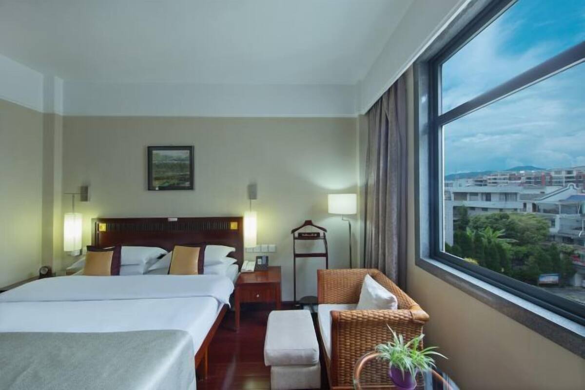 A hotel room with two beds, a large window with a city view, a wicker chair, and a small table.