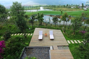 A wooden deck with two lounge chairs and a small table, surrounded by lush greenery and a view of a golf course and water features. - Landu Business Hotel (Tengchong)