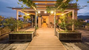 Um edifício moderno com deck de madeira que leva a uma entrada coberta, rodeado por floreiras e vegetação.